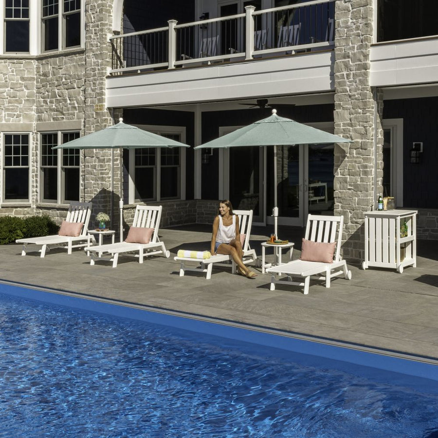 A woman sits on a POLYWOOD Vineyard 3-Piece Chaise with Wheels Set on a poolside patio by a stone house.