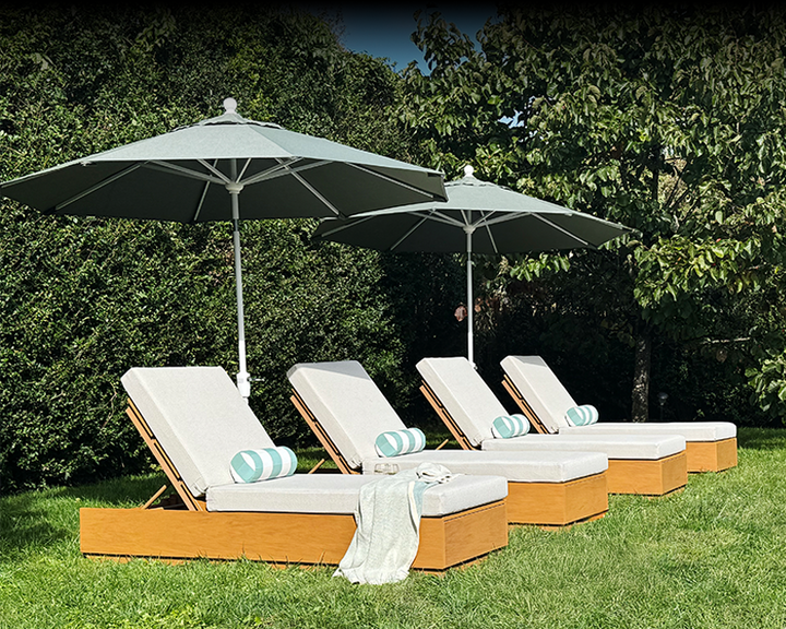 Four brown POLYWOOD chaise lounges with white cushions sit beneath umbrellas on a sunny lawn surrounded by tall hedges.