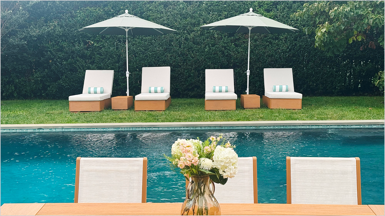 POLYWOOD chaise lounges and umbrellas sit on a lawn by a pool and tall hedges; chairs line a tan table that holds a vase of flowers in the foreground.