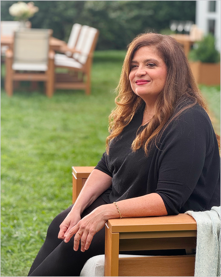 Chef Alex Guarnaschelli sits in a brown POLYWOOD deep seating chair on a lawn with more outdoor furniture behind her.