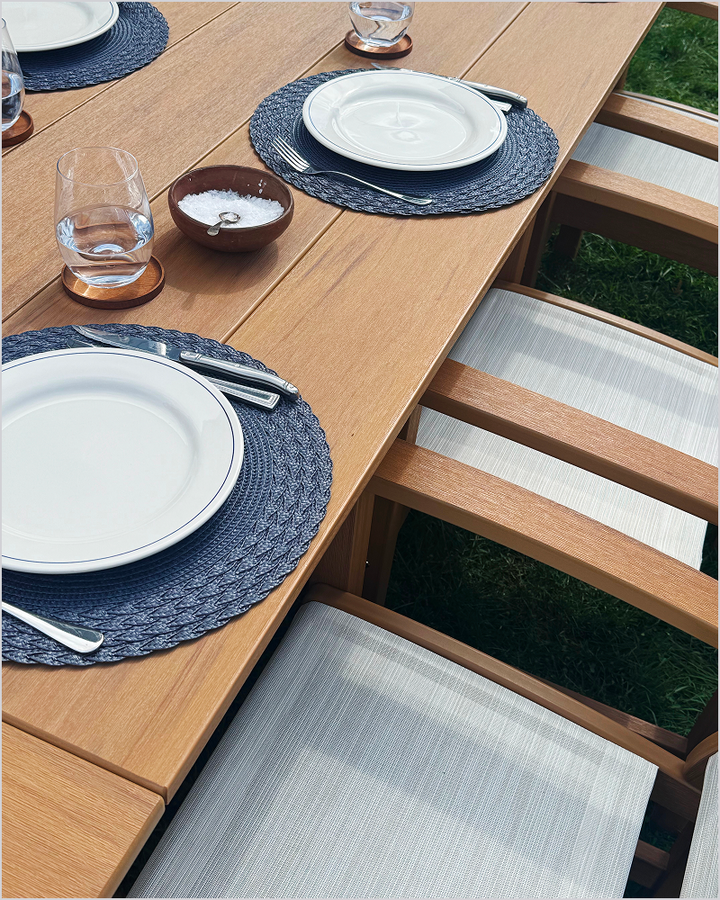 A brown POLYWOOD dining table is set with plates, glasses, and dark woven placemats by dining chairs with white sling seats.