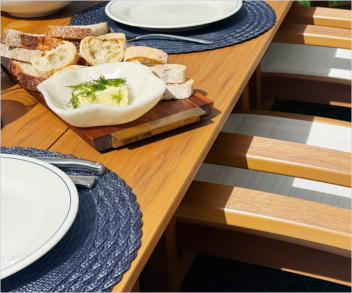 A brown POLYWOOD dining table holds a wooden tray with sliced bread and butter, plates, silverware, and woven placemats.