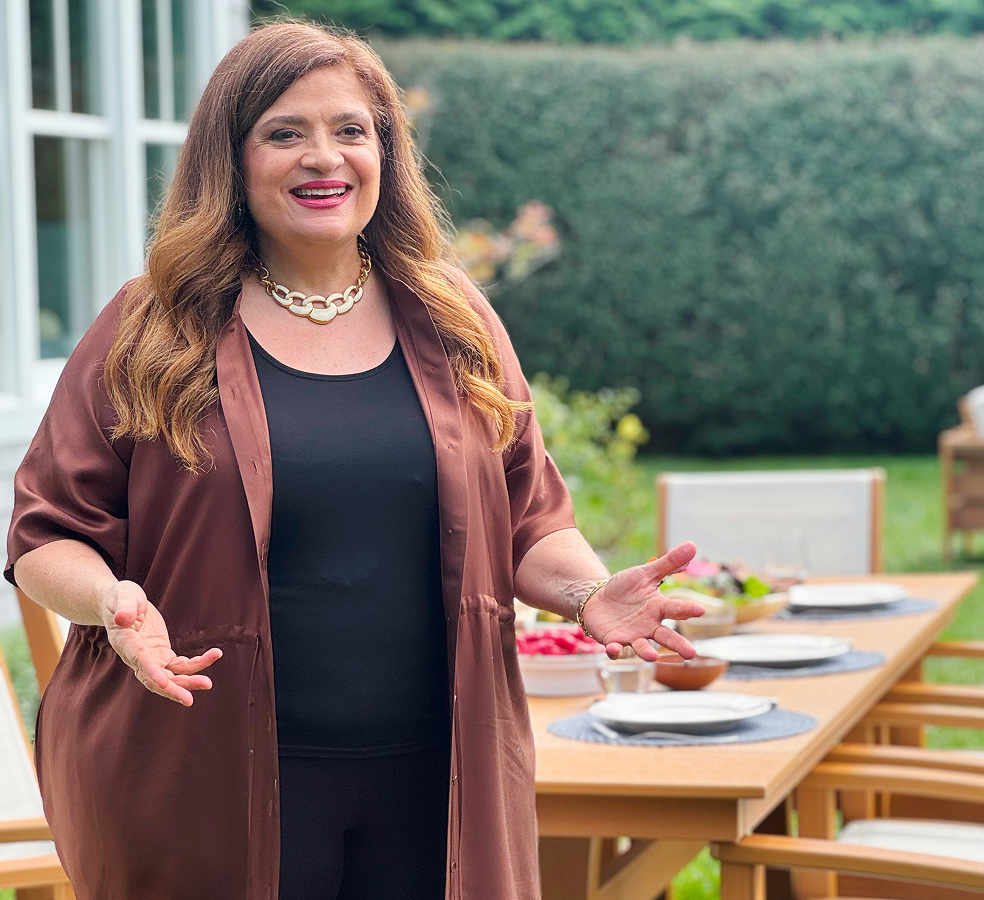 Chef Alex Guarnaschelli stands before a tan POLYWOOD dining set in a backyard setting; the table is set for a meal.