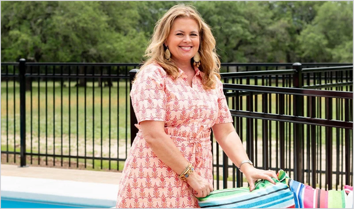Designer Jessica Duce stands outdoors by a pool wearing a pink and coral pineapple-print dress and holding a striped beach towel. A black metal fence and green trees are in the background.